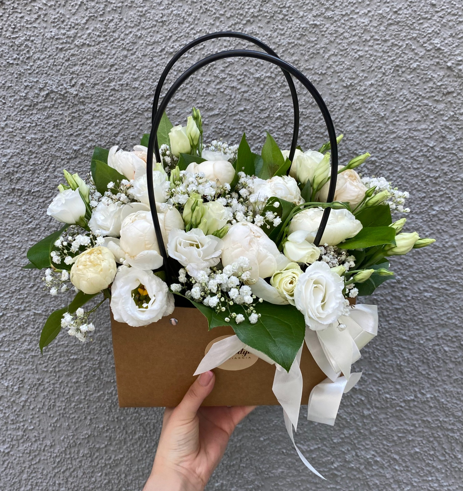 Elegancki kosz prezentowy z białych róż, eustomy i gipsówki, umieszczony w brązowej torbie z czarnymi uchwytami i ozdobiony białą wstążką, trzymany na tle szarej ściany o chropowatej fakturze.