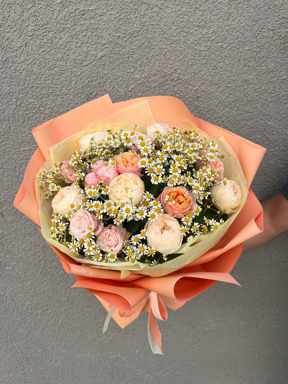 Bukiet pastelowych róż i drobnych białych margaretek, owinięty w pomarańczowy i żółty papier, trzymany na tle szarej ściany.
