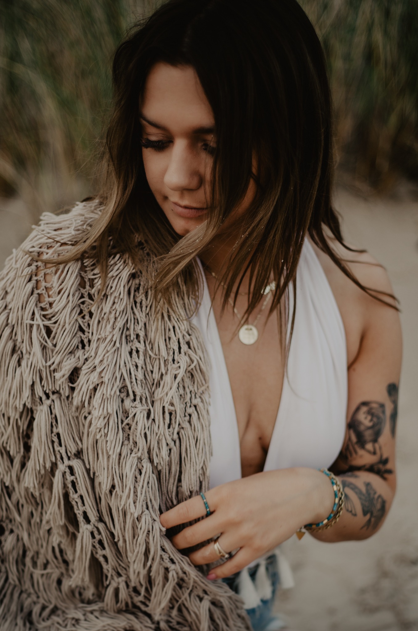 Portret kobiety w białej sukience i narzutce z frędzlami na plaży w Ustce, z tatuażem na ramieniu i biżuterią, spoglądającej w dół.