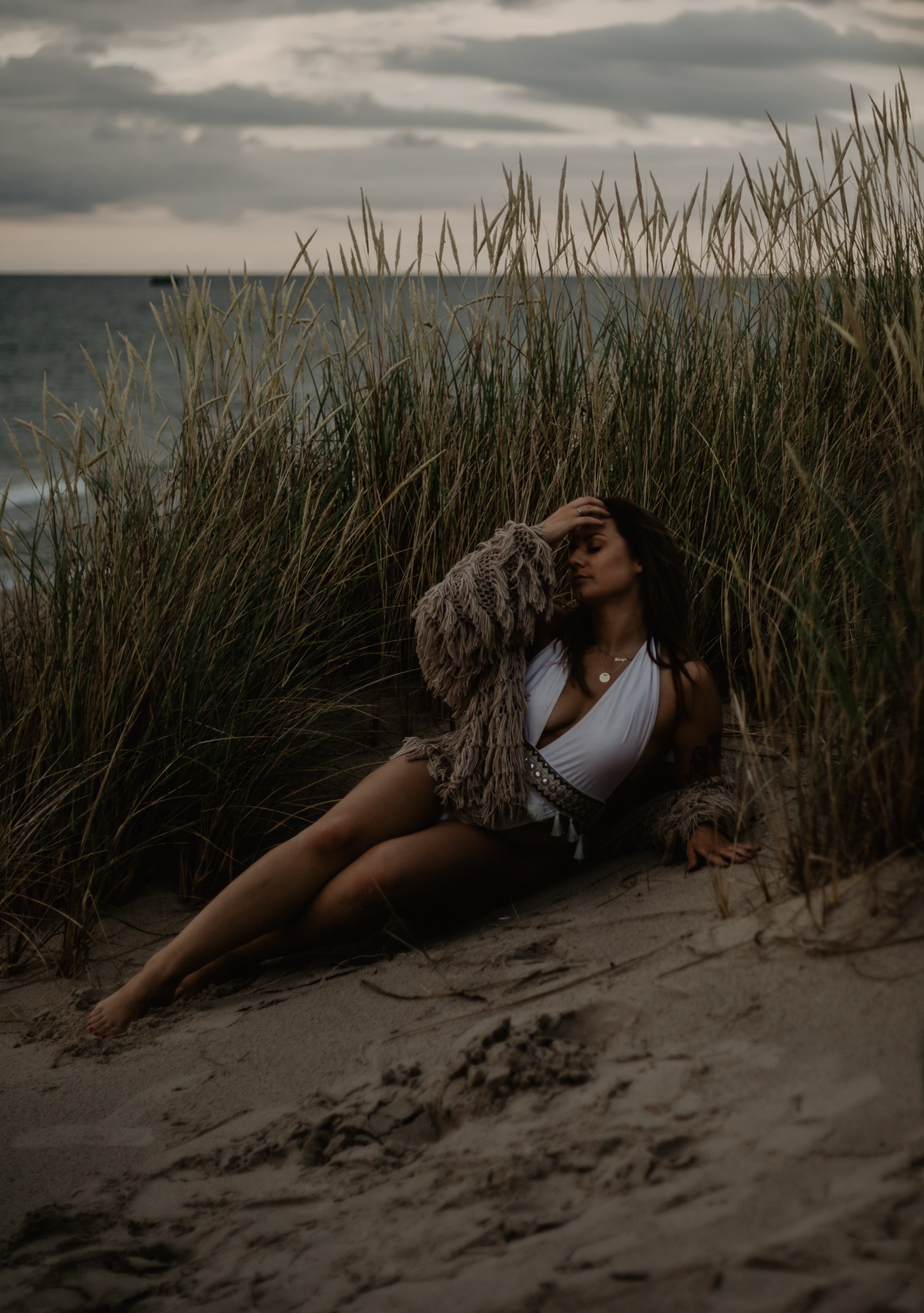 Kobieta w białym stroju kąpielowym i narzutce z frędzlami pozuje leżąc na piaszczystej plaży w Ustce, otoczona trawą, z widokiem na morze i pochmurne niebo w tle.
