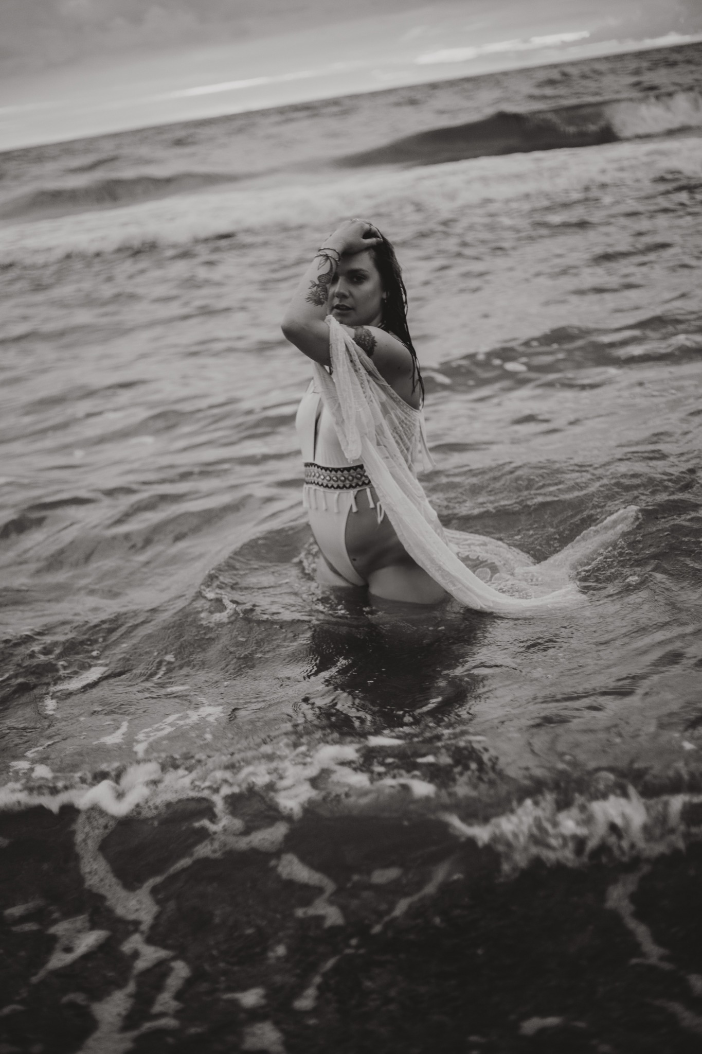Czarno-biała fotografia kobiety w stroju kąpielowym i narzutce, stojącej w morzu z ręką na głowie, widoczne tatuaże na ramieniu, w tle wzburzone fale i pochmurne niebo.
