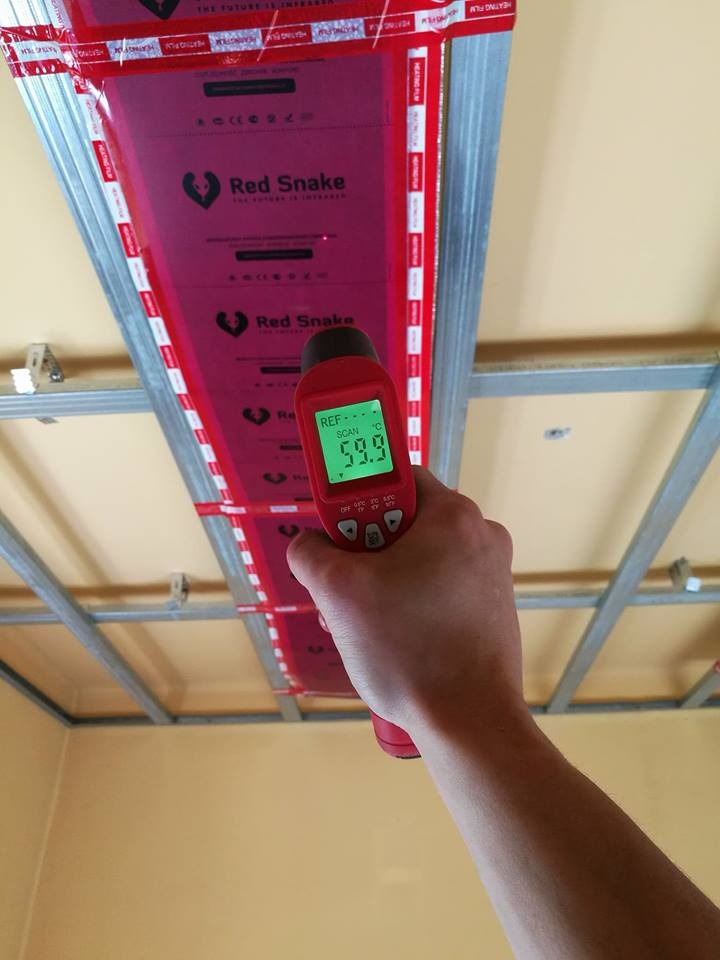 Pomiar temperatury folii grzewczej Red Snake na suficie podwieszanym z użyciem termometru laserowego, wskazującego 59.9 stopni Celsjusza.