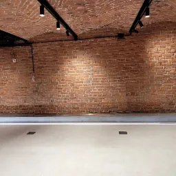 Wnętrze piwnicy z ceglanym sklepieniem, oświetleniem punktowym na czarnych szynach i jasną, gładką posadzką z listwą LED przy ścianie. Widoczna instalacja elektryczna natynkowa i szary filar.