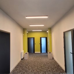 Premium Bud - Długi korytarz biurowy z szarą wykładziną w geometryczne wzory, podłużnymi lampami LED i ścianami w odcieniach bieli i limonki, z widocznymi drzwiami po obu stronach i grzejnikiem przy ścianie.