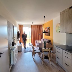 Premium Bud - Widok na wyremontowane mieszkanie z aneksem kuchennym, widoczny pracownik w ubraniu roboczym wchodzący do salonu, okrągłe lustra na ścianie, drewniane szafki kuchenne i stół z krzesłami.