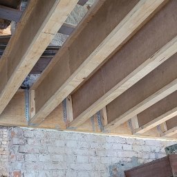 Premium Bud - Widok z dołu na konstrukcję drewnianego stropu z płytami OSB, przymocowaną do starej, ceglanej ściany za pomocą metalowych łączników; widoczne fragmenty starego stropu.