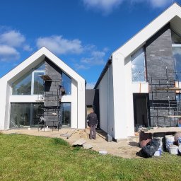 Premium Bud - Dwa nowoczesne domy w trakcie budowy, białe elewacje, czarne elementy dekoracyjne, rusztowania, pracownicy w trakcie pracy, widoczne puszki z farbą, trawnik przed domami, błękitne niebo z chmurami.