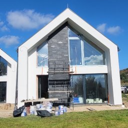 Premium Bud - Nowoczesny dom w trakcie budowy z białymi ścianami i fragmentem elewacji wykończonym ciemną cegłą, widoczne rusztowanie i puszki z farbą na trawniku.