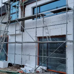 Dom w trakcie ocieplania styropianem, widoczne rusztowania, okna z folią ochronną i stos styropianu na ziemi.