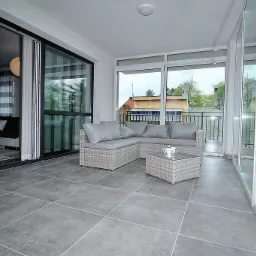 Zadaszony taras z szarymi płytkami podłogowymi, rattanowym narożnikiem z szarymi poduszkami i stolikiem kawowym, widok na zewnątrz przez przesuwne szklane drzwi i okna.