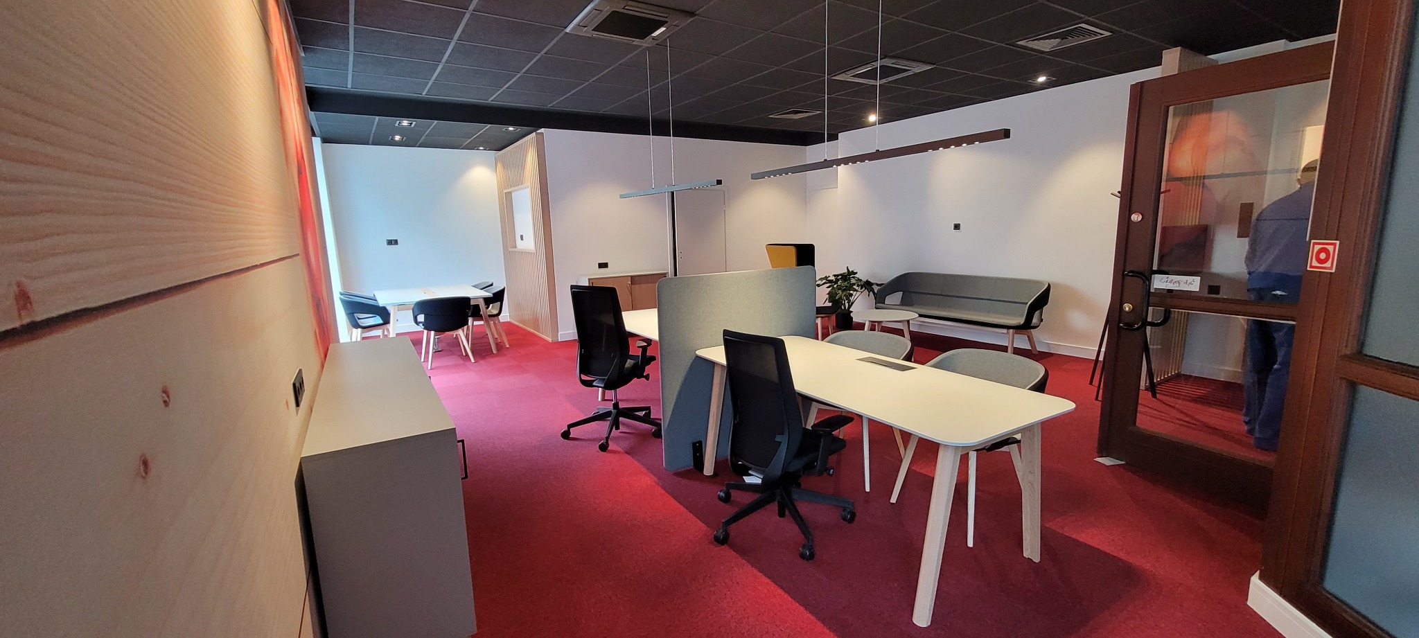 Nowoczesne biuro z czerwonym dywanem, białymi biurkami z drewnianymi nogami, szarą sofą i ścianą z imitacją drewna, widziane z perspektywy wejścia.