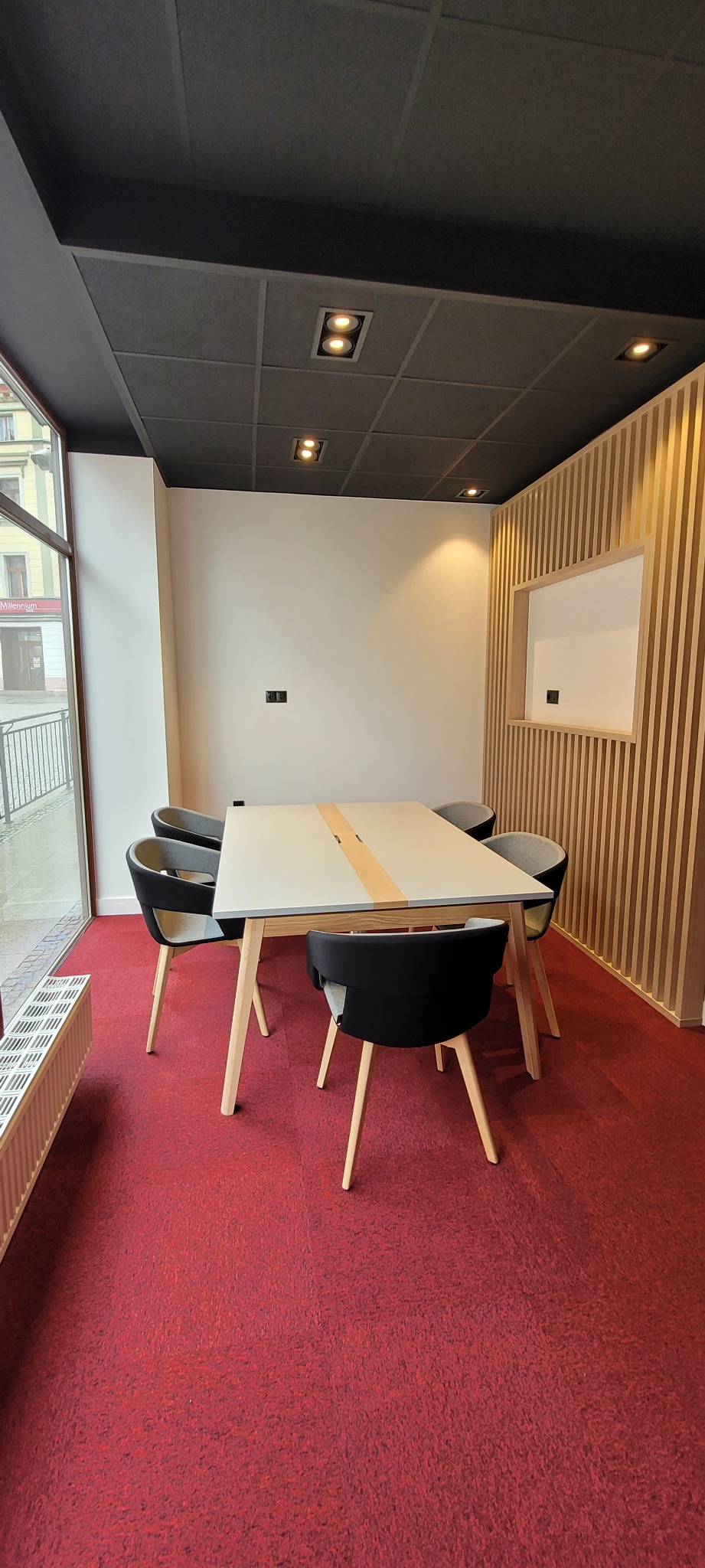 Nowoczesne biuro z czerwonym dywanem, jasnym stołem konferencyjnym i krzesłami z czarnym oparciem, oświetlenie sufitowe punktowe, ściana z drewnianych paneli pionowych.