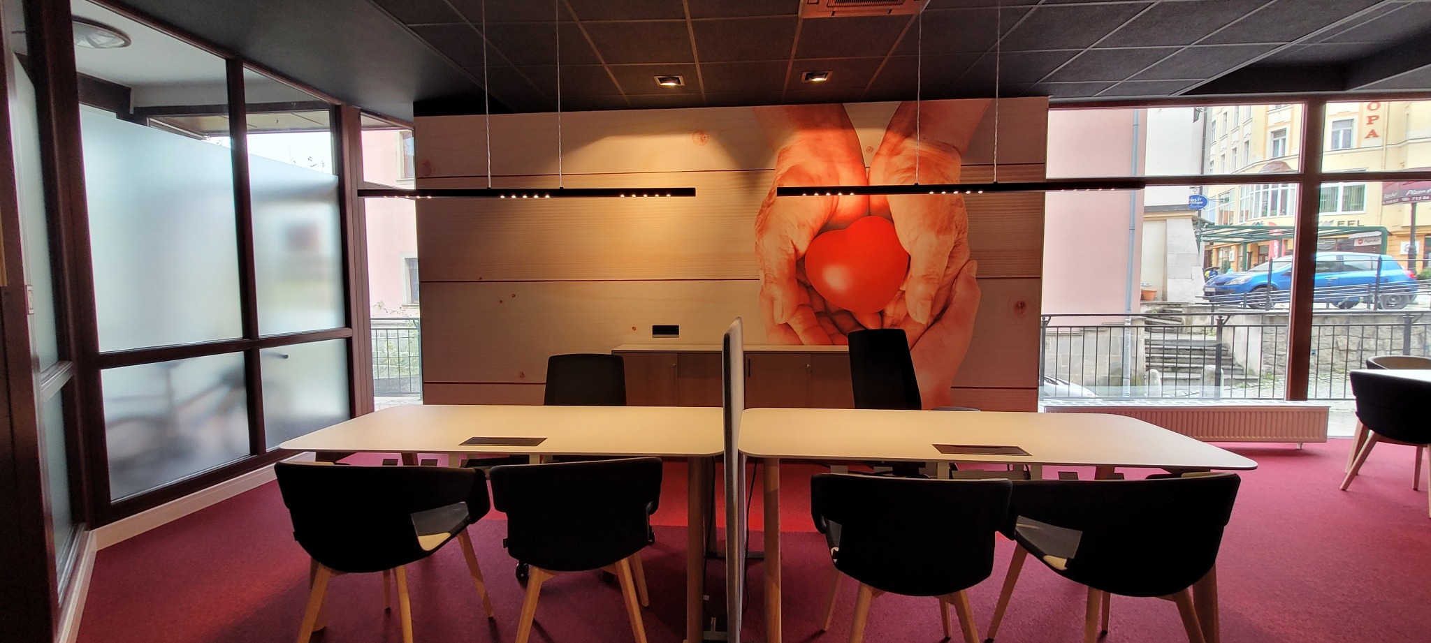 Nowoczesne biuro z białymi stołami, czarnymi krzesłami na drewnianych nogach, czerwonym dywanem i ścianą z grafiką dłoni trzymających czerwone serce, oświetlenie liniowe LED