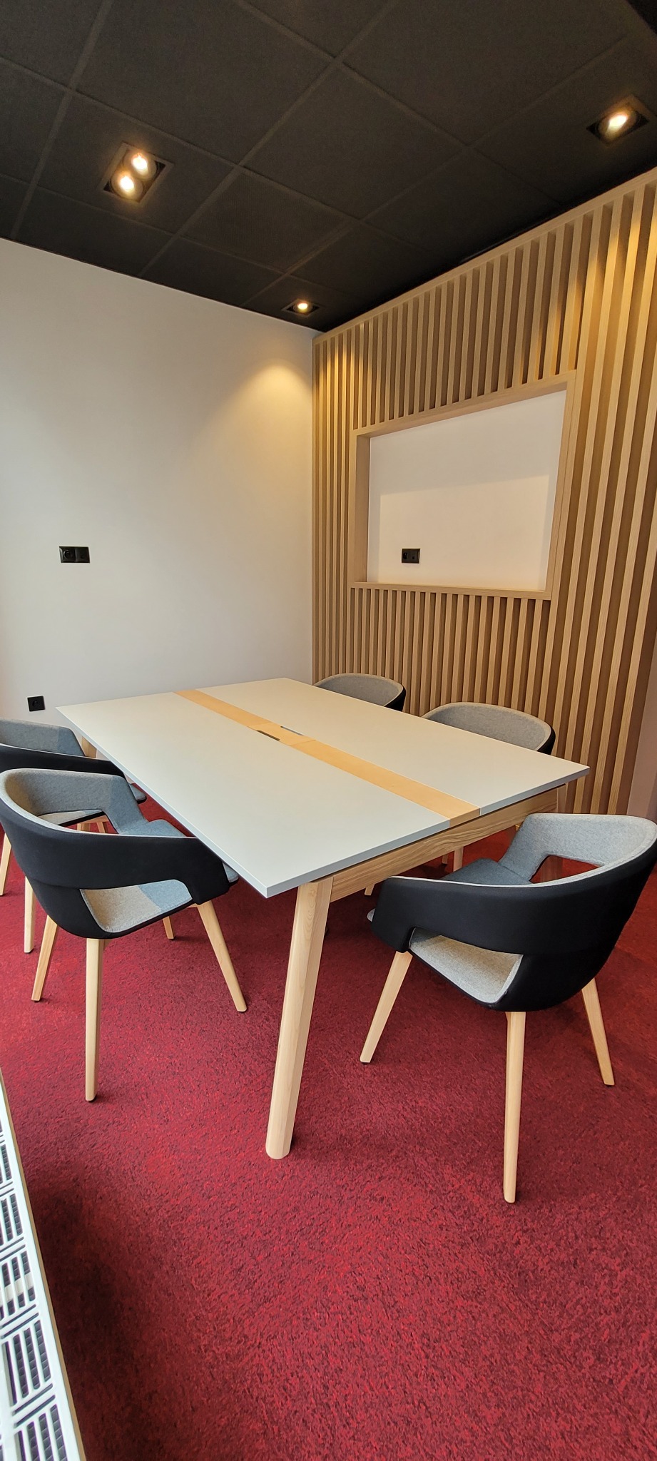 Nowoczesna sala konferencyjna z szarym stołem z drewnianymi nogami, czarnymi krzesłami z szarą tapicerką, czerwoną wykładziną podłogową i dekoracyjną ścianą z drewnianych listew.