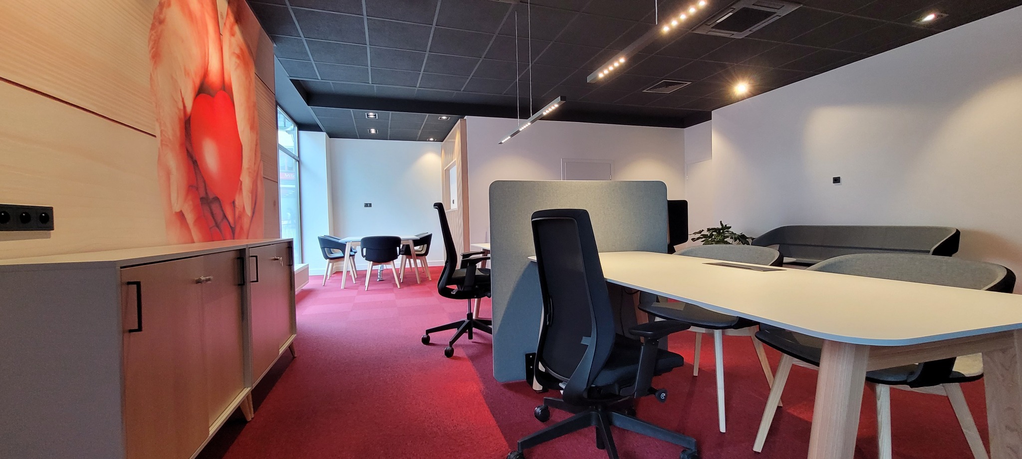 Nowoczesne biuro z czerwonym dywanem, jasnymi biurkami i czarnymi krzesłami, uzupełnione szafką z grafiką na ścianie.