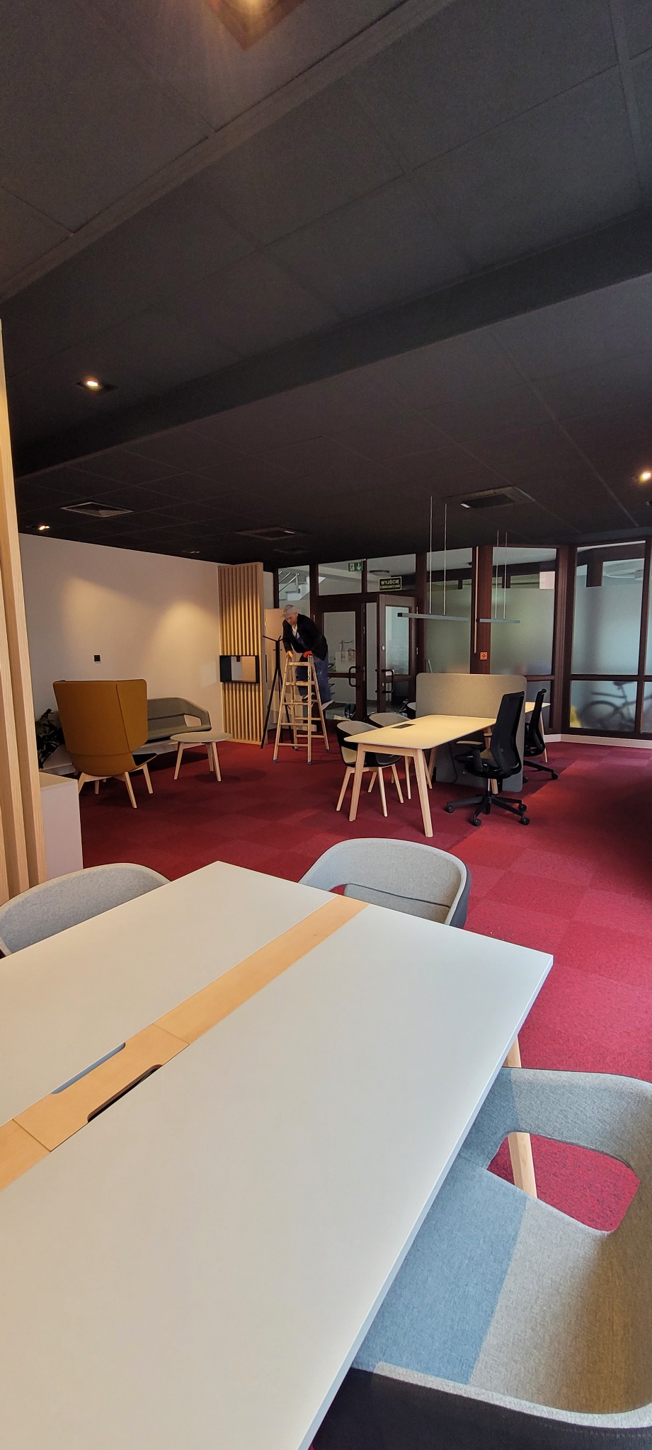 Pracownik na drabinie w biurze z ciemnym sufitem, czerwonym dywanem i jasnymi meblami, prawdopodobnie podczas prac wykończeniowych lub drobnych napraw.