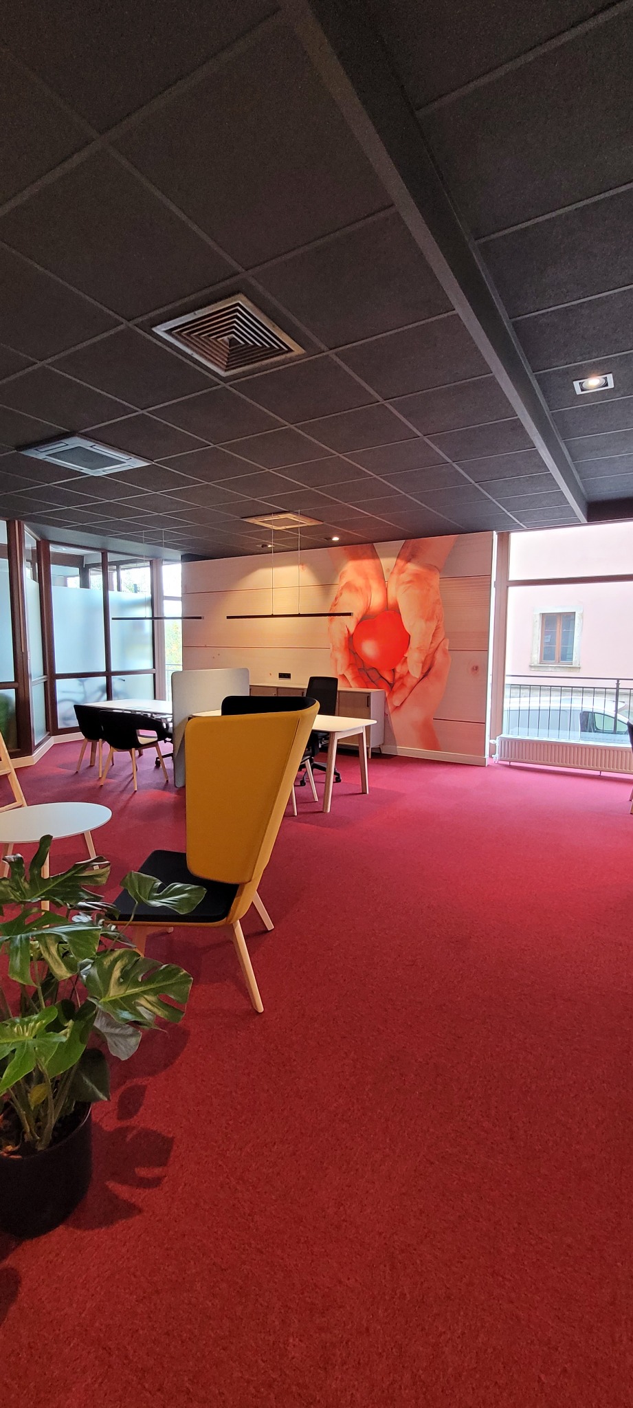 Nowoczesne biuro z czerwoną wykładziną dywanową, żółtym krzesłem i dekoracyjną fototapetą z motywem dłoni trzymających serce.