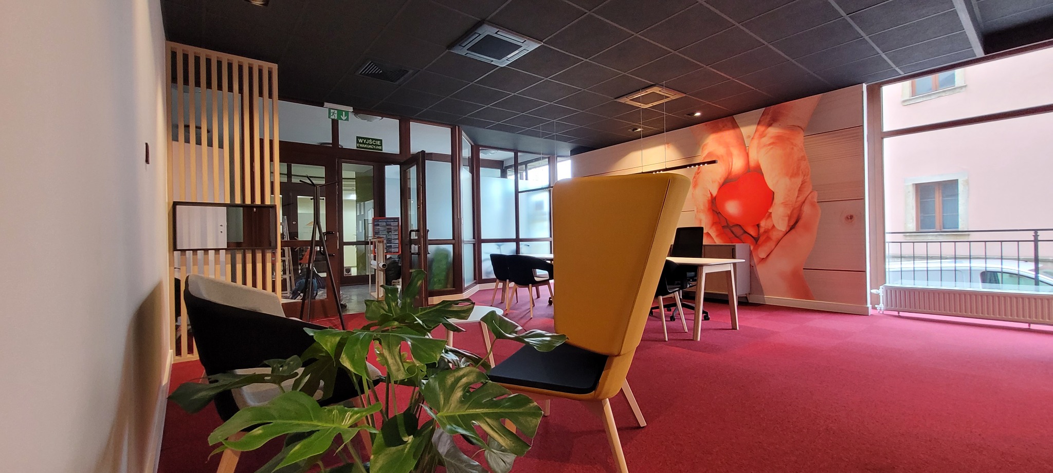 Nowoczesne biuro z czerwonym dywanem, żółtym fotelem z wysokim oparciem, rośliną doniczkową i grafiką na ścianie przedstawiającą dłonie trzymające czerwone serce.