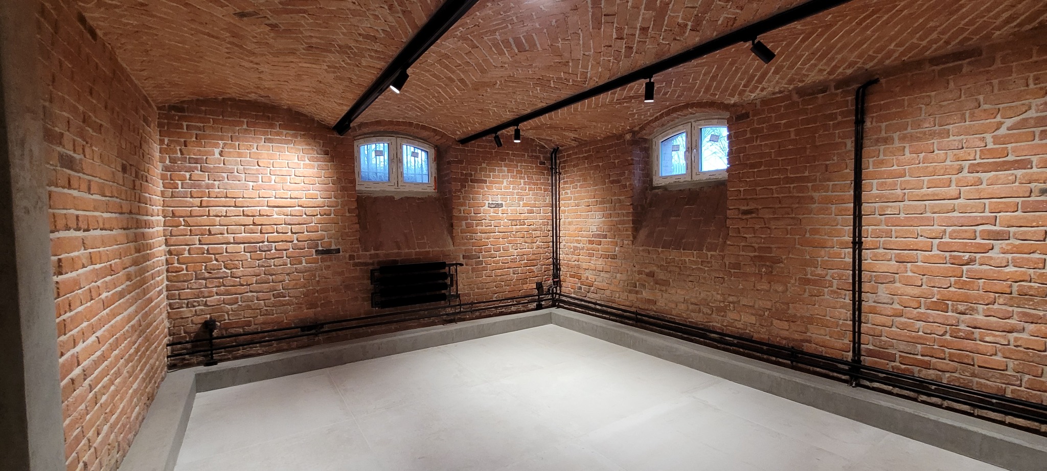 Piwnica z ceglanym sklepieniem łukowym, oświetleniem punktowym, dwoma małymi oknami i szarymi płytkami podłogowymi.