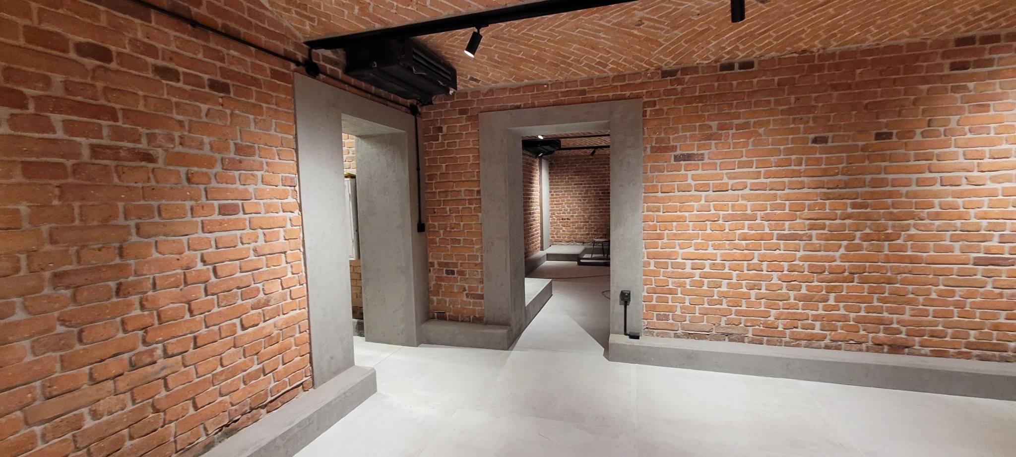 Korytarz z ceglanymi ścianami i sklepionym sufitem, oświetlony reflektorami punktowymi, z szarymi, betonowymi ościeżnicami drzwi i jasną, gładką podłogą.