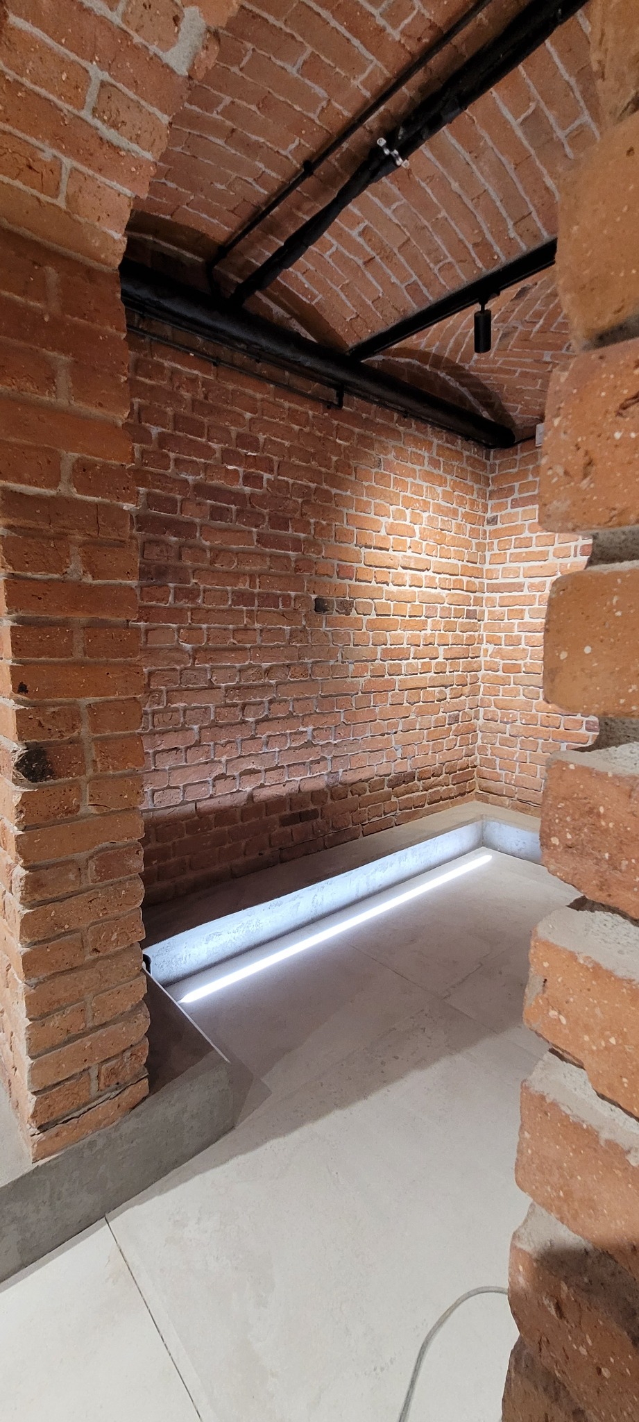 Klimatyczne wnętrze z ceglanym sklepieniem łukowym, oświetlone listwą LED wzdłuż podłogi i czarnymi rurami pod sufitem, ukazujące surowy, industrialny charakter.