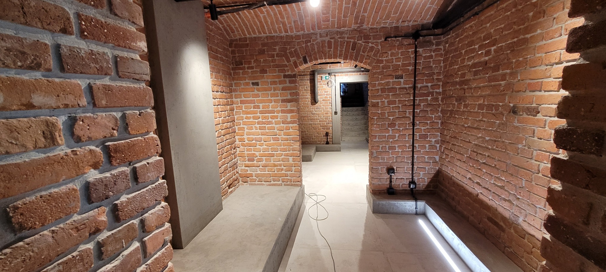 Korytarz z ceglanymi ścianami i sklepieniem łukowym, oświetlony podłogowymi lampami LED, z widocznymi elementami instalacji elektrycznej i betonowymi wstawkami.
