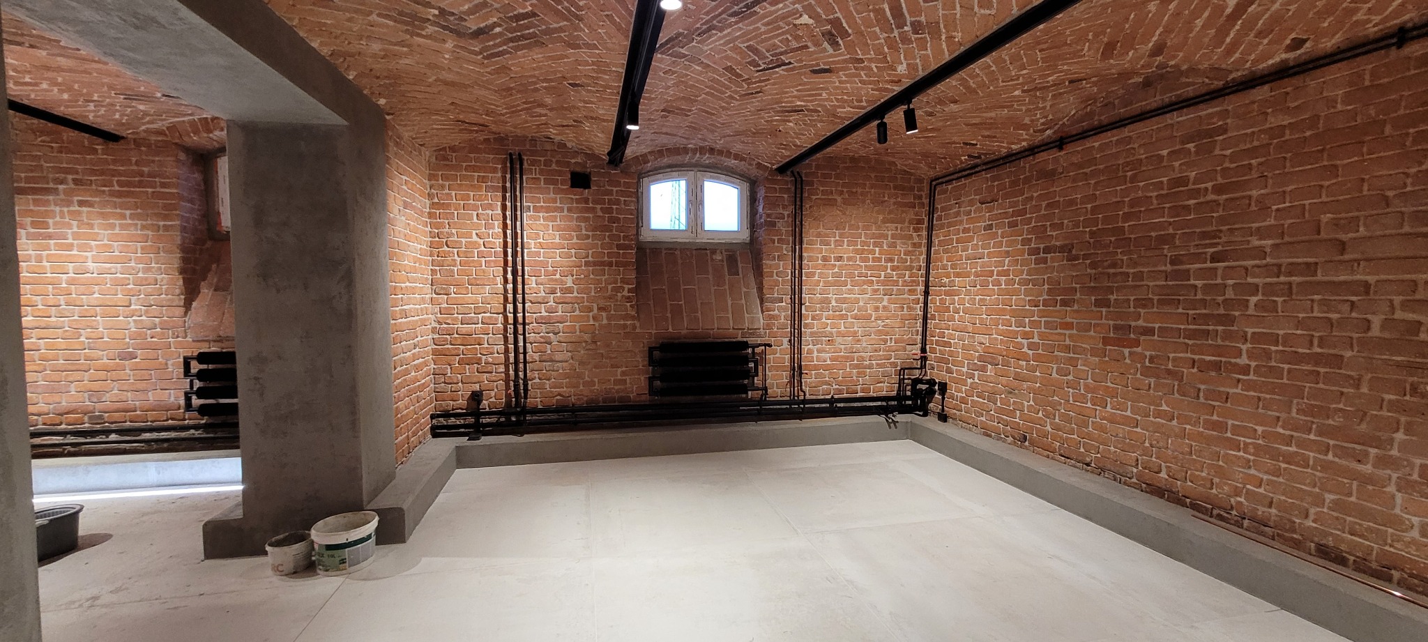 Wnętrze z ceglanym sklepieniem i ścianami, widoczne rury grzewcze, grzejnik żeberkowy pod oknem i świeżo wylana, jasna posadzka. Dwa wiadra z farbą w lewym dolnym rogu sugerują prace wykończeniowe.