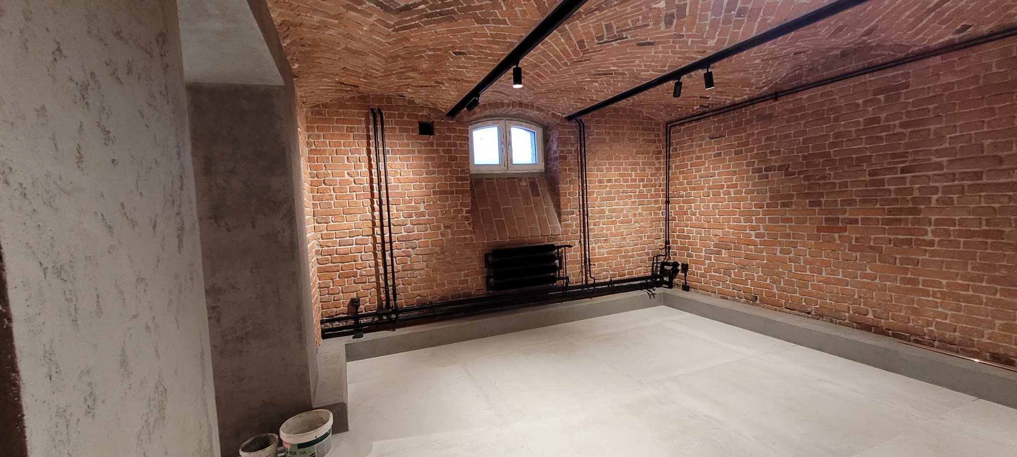 Wykończone wnętrze z ceglanym sklepieniem, czarnym oświetleniem szynowym, grzejnikiem żeliwnym i jasną podłogą z gresu. Widoczne rury instalacji biegnące wzdłuż ścian. Ściana po lewej stronie...