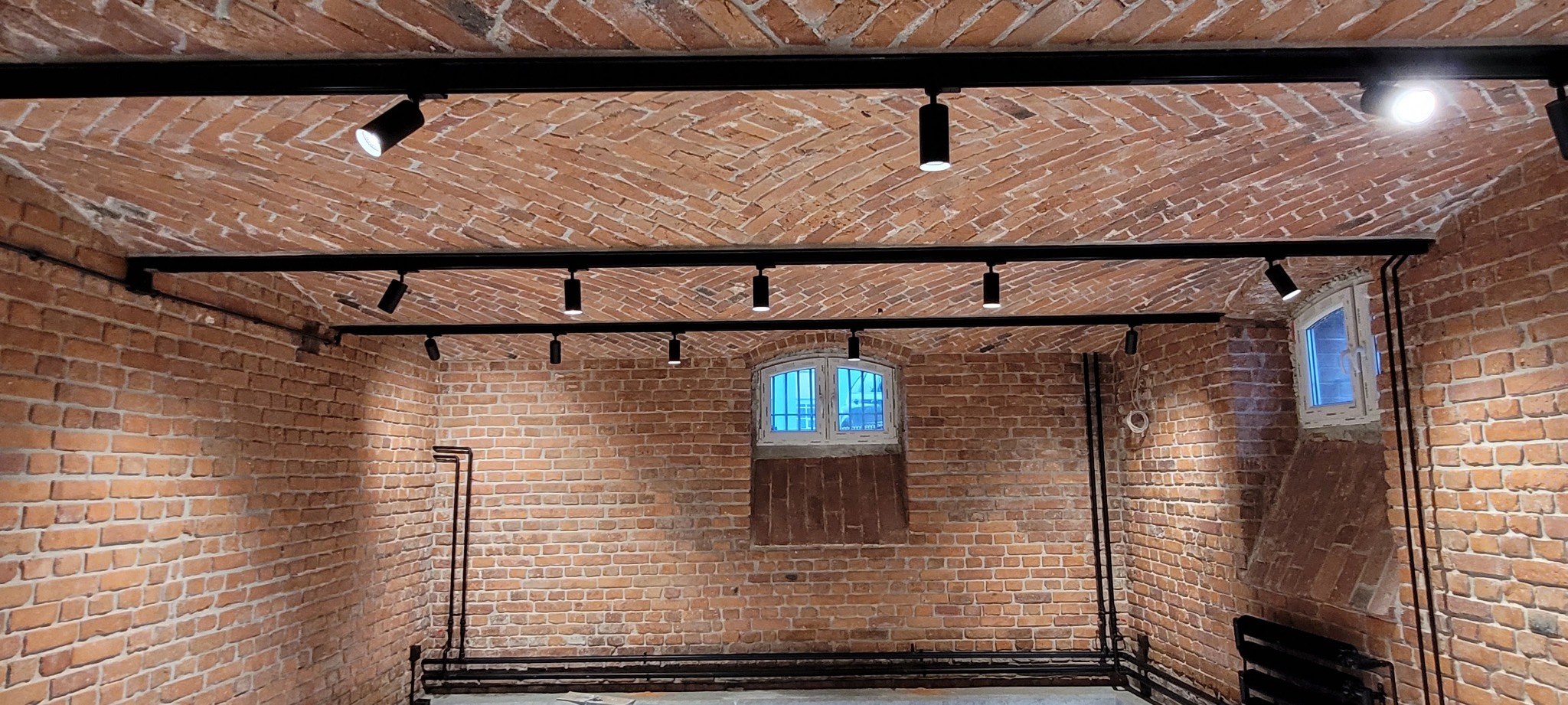 Wnętrze piwnicy z ceglanym sklepieniem łukowym, oświetlone czarnymi reflektorami na szynoprzewodach. Widoczne okno z niebieskim światłem dziennym i instalacje elektryczne na ścianach.