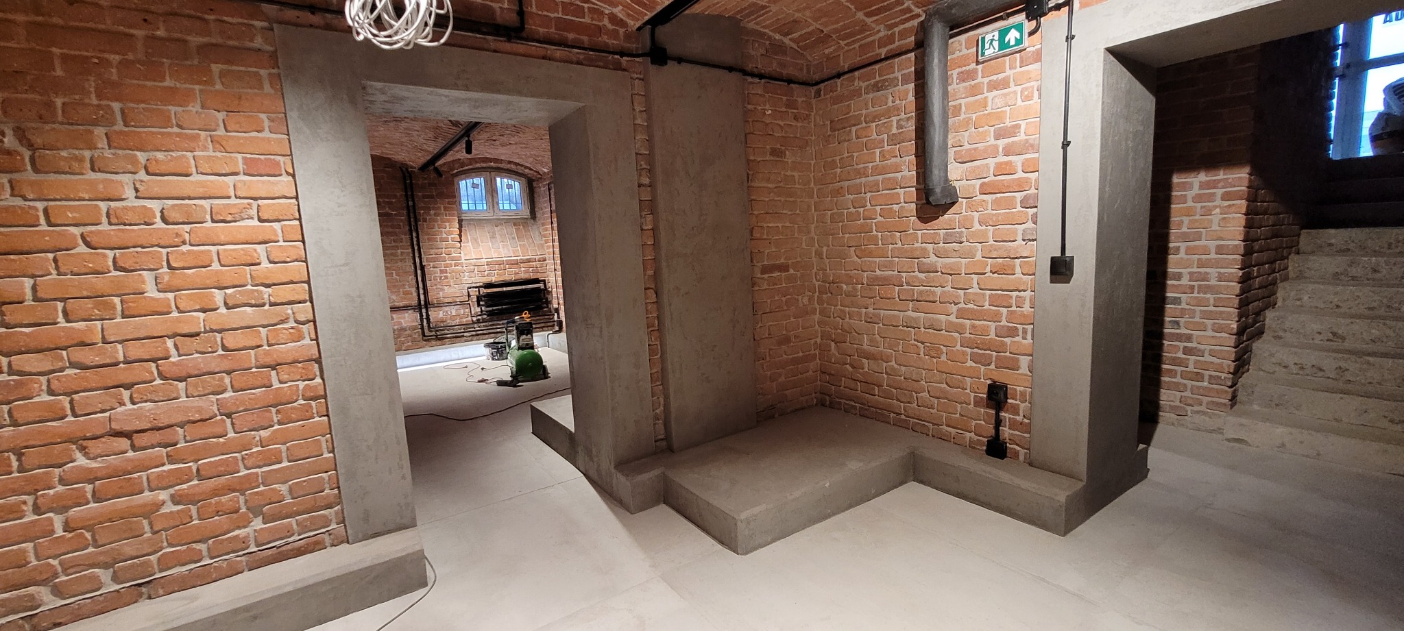 Wnętrze piwnicy po renowacji z ceglanymi ścianami, betonowymi filarami i schodami, widoczne instalacje elektryczne i wentylacyjne, w tle kompresor malarski.