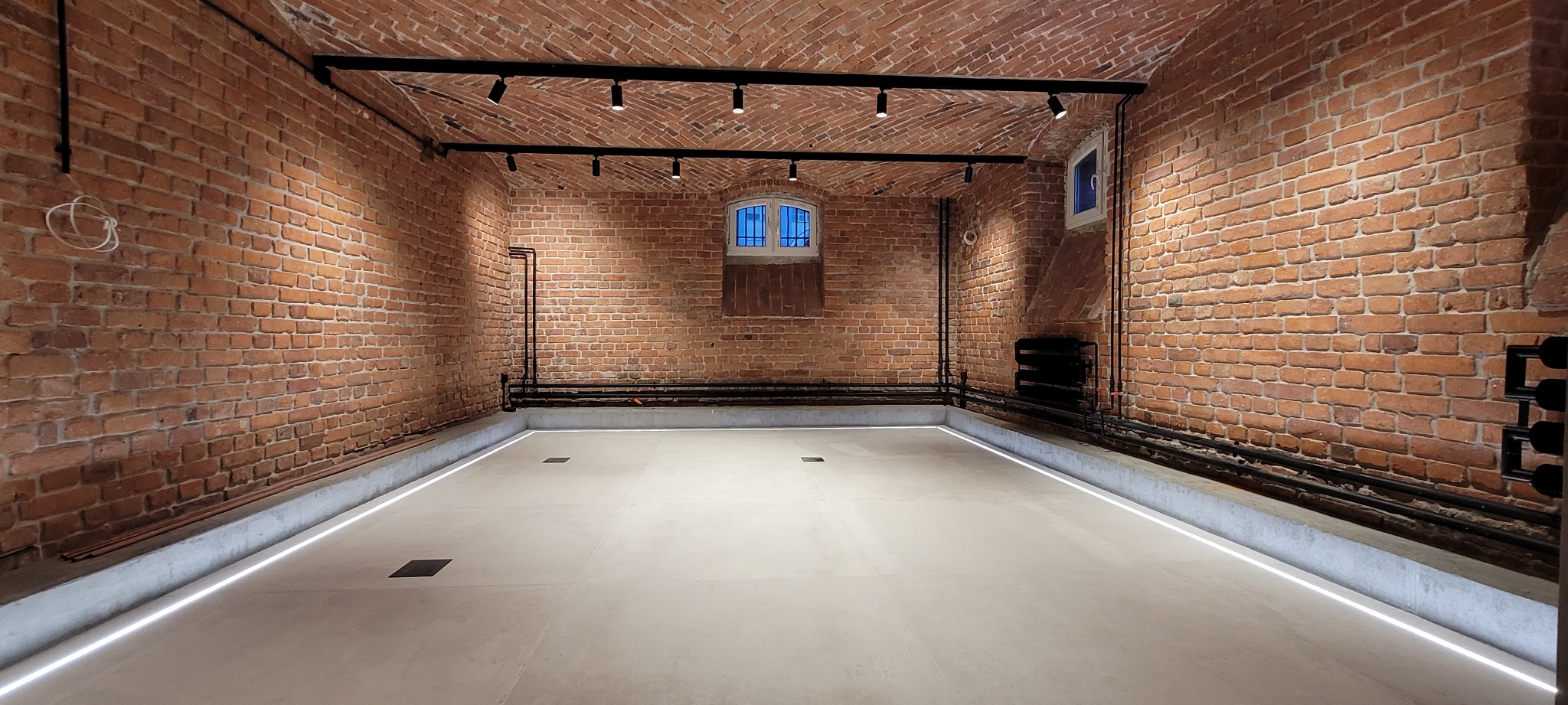 Klimatyczne wnętrze z ceglanymi ścianami i sklepieniem, oświetlone listwami LED wzdłuż podłogi i reflektorami na szynoprzewodzie, z widocznymi rurami instalacji i małym oknem z okiennicą.