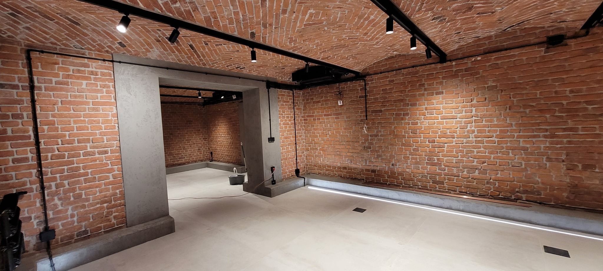 Surowe wnętrze z ceglanymi ścianami i sklepieniem, oświetlone czarnymi reflektorami na szynoprzewodach, z betonowymi filarami i jasną podłogą, w tle widoczne wiadro i narzędzia.