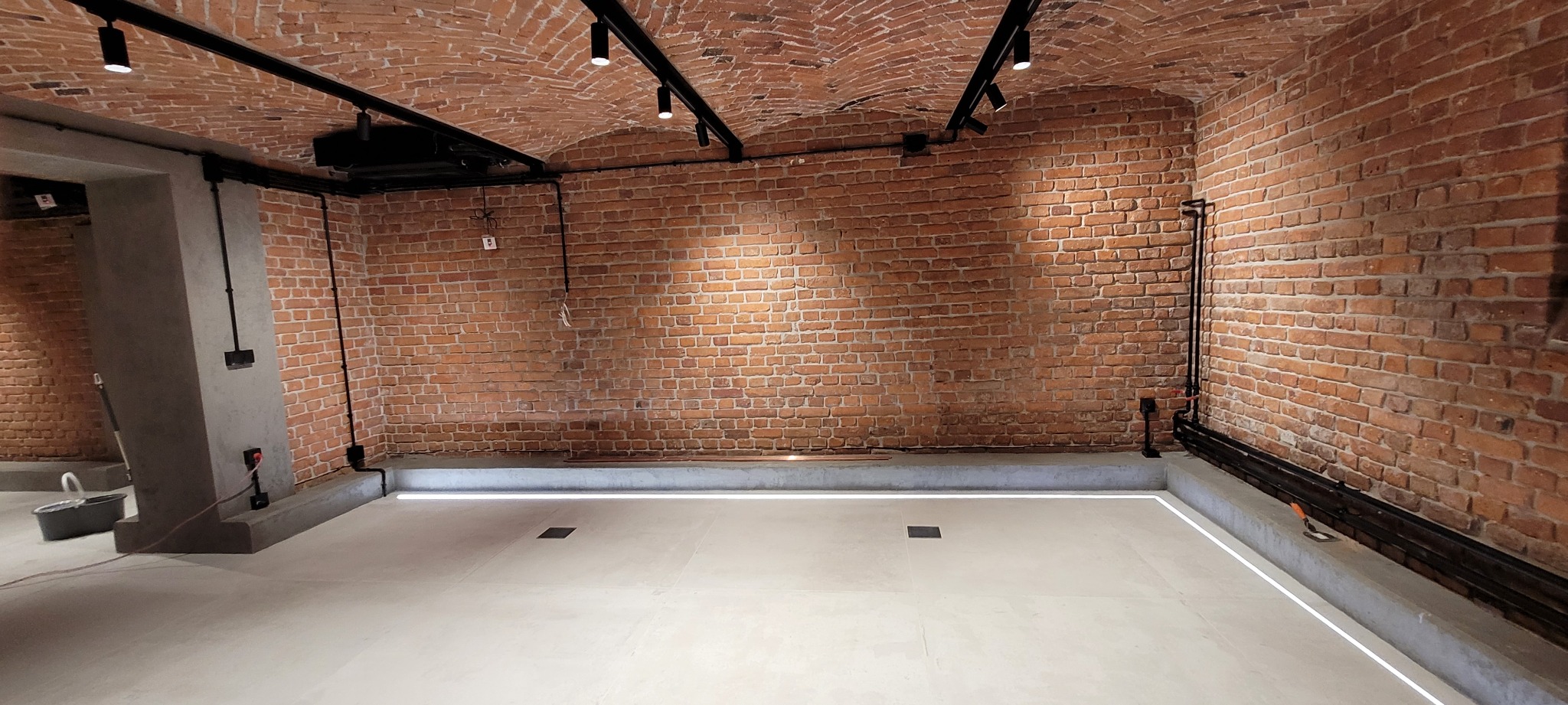 Wnętrze piwnicy z ceglanym sklepieniem, oświetleniem punktowym na czarnych szynach i jasną, gładką posadzką z listwą LED przy ścianie. Widoczna instalacja elektryczna natynkowa i szary filar.