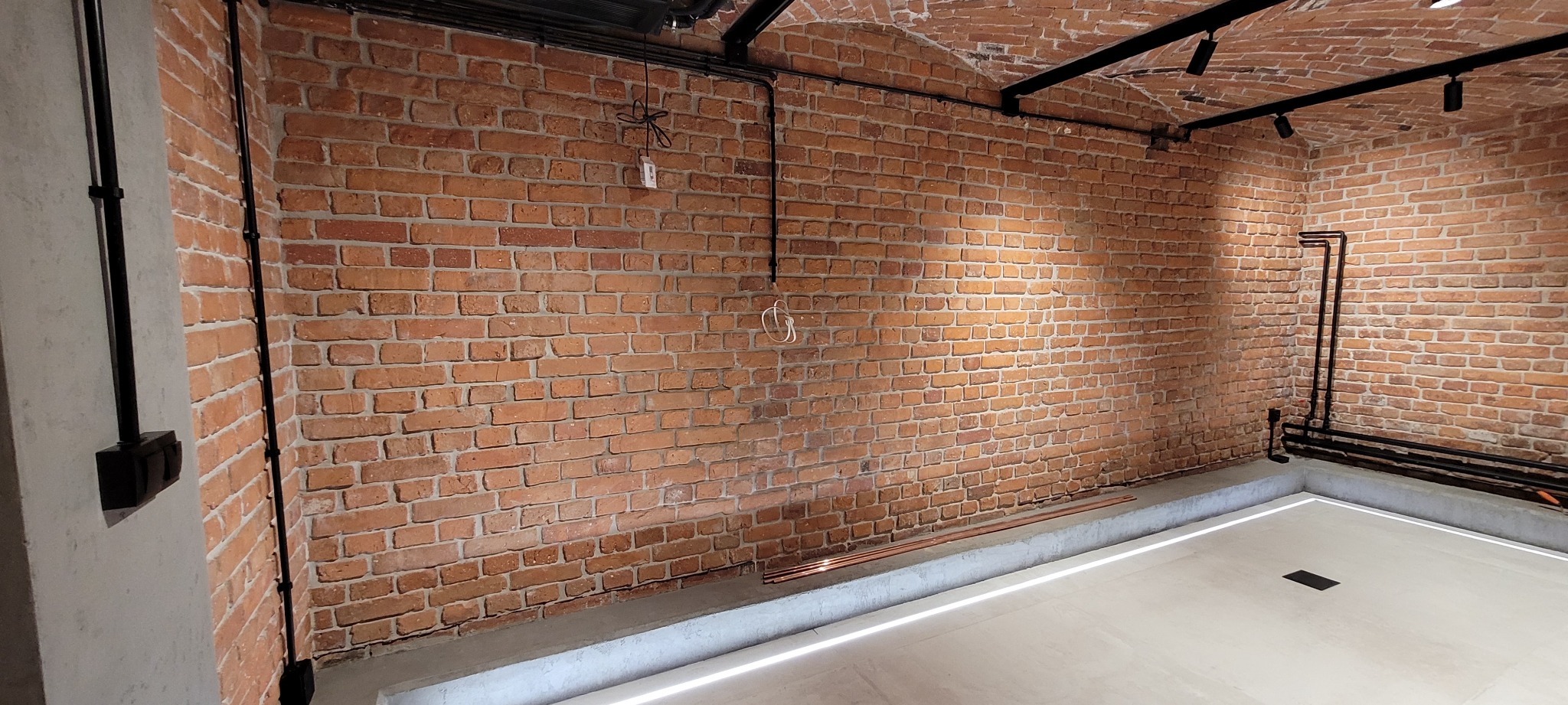 Wnętrze z surową ceglaną ścianą, widoczne instalacje elektryczne, rury oraz podświetlana podłoga, tworzące nowoczesny loftowy charakter.