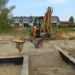 Przygotowanie terenu pod fundamenty: ubijarki wibracyjne zagęszczają piasek w wykopie, w tle żółta koparko-ładowarka i zabudowa szeregowa.