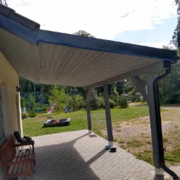 Zadaszony taras z drewnianą konstrukcją, rynną i widokiem na ogród z huśtawkami, rowerem i psem śpiącym na krześle.