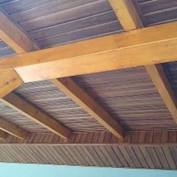 Drewniana konstrukcja dachu z widocznymi belkami i podbitką w odcieniach brązu, oświetlona wiszącą żarówką.