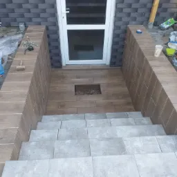 Wykończone płytkami schody wejściowe do domu z drewnopodobną podłogą i szarą elewacją z cegły klinkierowej, widoczne narzędzia i materiały budowlane.