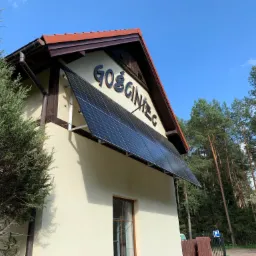 Panel słoneczny zamontowany nad oknem budynku z napisem 'Gościniec', widok z dołu na błękitne niebo