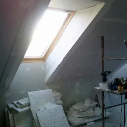 Poddasze w trakcie adaptacji z widocznym oknem dachowym, stertą płyt izolacyjnych z czarnymi kropkami i prowizorycznym stołem roboczym z narzędziami, ściany przygotowane pod wykończenie.