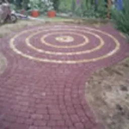 Ułożona z czerwonej kostki brukowej ścieżka w ogrodzie, tworząca dekoracyjny wzór koncentrycznych okręgów z żółtymi akcentami. Widoczny fragment trawnika i niewielkie drzewko. Część kostki jeszcze...