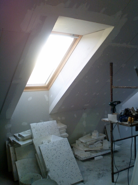 Poddasze w trakcie adaptacji z widocznym oknem dachowym, stertą płyt izolacyjnych z czarnymi kropkami i prowizorycznym stołem roboczym z narzędziami, ściany przygotowane pod wykończenie.