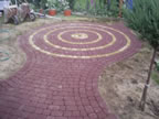 Ułożona z czerwonej kostki brukowej ścieżka w ogrodzie, tworząca dekoracyjny wzór koncentrycznych okręgów z żółtymi akcentami. Widoczny fragment trawnika i niewielkie drzewko. Część kostki jeszcze...