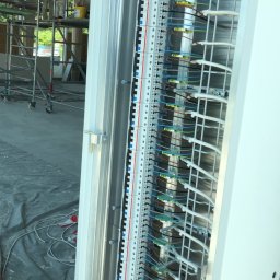 EL-TOM - Otwarta skrzynka rozdzielcza z widocznymi listwami zaciskowymi, przewodami i elementami montażowymi, w tle rusztowanie i pracownicy budowlani.