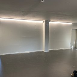 EL-TOM - Wnętrze po remoncie: świeżo pomalowane jasne ściany, szara podłoga, zamontowane oświetlenie liniowe LED pod sufitem, widoczne dwa wiadra malarskie w prawym dolnym rogu.