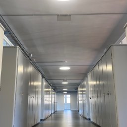 EL-TOM - Długi korytarz z rzędami białych szafek po obu stronach, oświetlony okrągłymi lampami sufitowymi i światłem dziennym wpadającym przez okno na końcu korytarza, widoczne instalacje elektryczne...