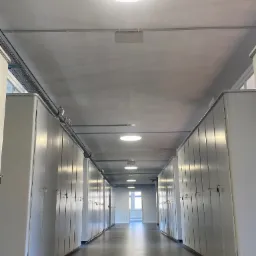 Długi korytarz z rzędami białych szafek po obu stronach, oświetlony okrągłymi lampami sufitowymi i światłem dziennym wpadającym przez okno na końcu korytarza, widoczne instalacje elektryczne...