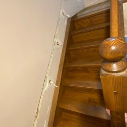 EL-TOM - Wnętrze domu w trakcie remontu: drewniane schody z balustradą, różowa ściana z bruzdami na przewody elektryczne i wystającymi przewodami, widoczne ślady gipsu.