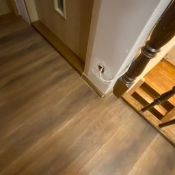 EL-TOM - Biały kabel wystający z dziury w ścianie przy schodach i drewnianej podłodze; widoczna stopa w skarpetce.