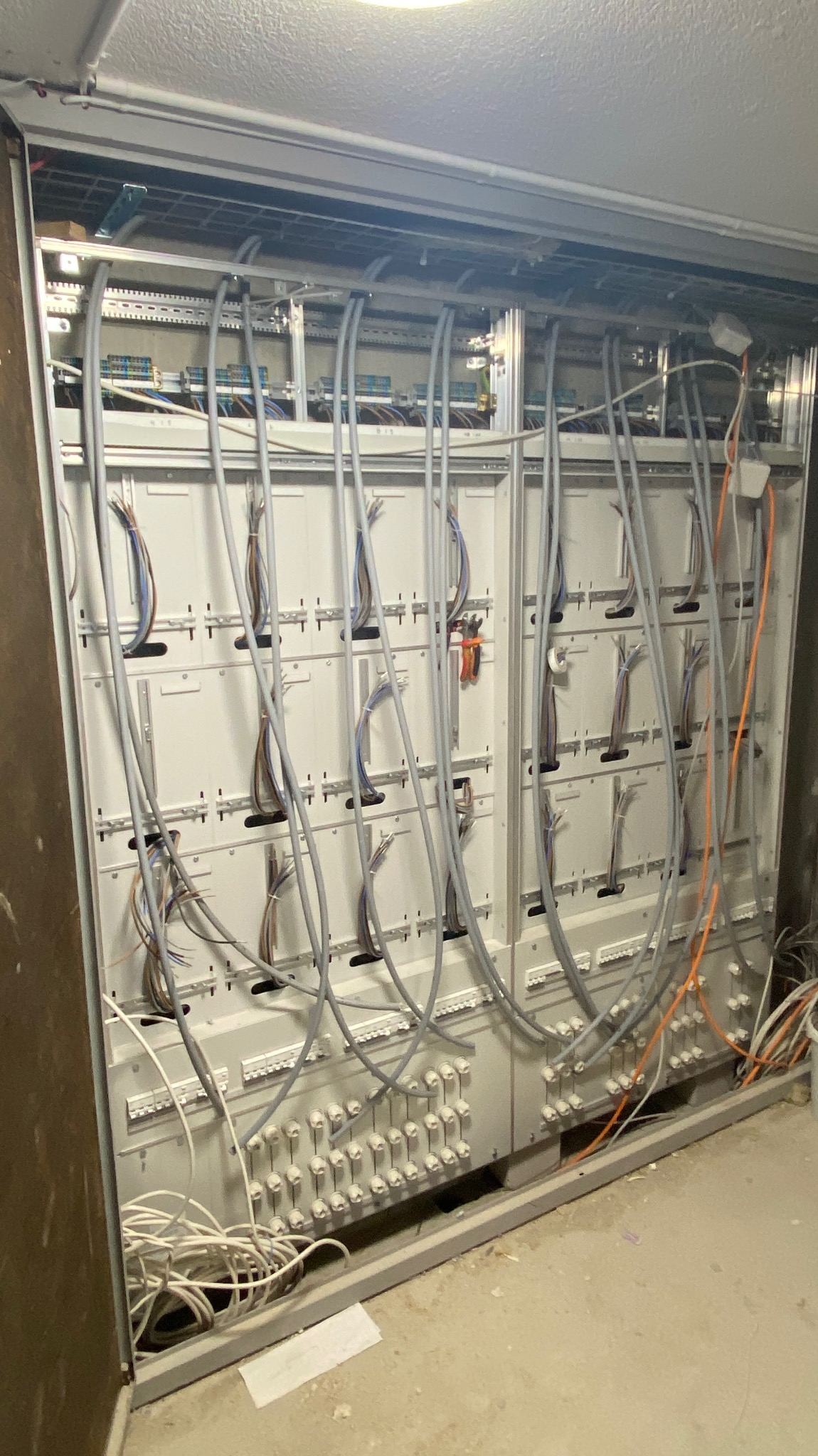 Rozdzielnica elektryczna w trakcie montażu z licznymi przewodami szarymi i pomarańczowymi, widoczne złącza i listwy montażowe, surowy stan wykończenia pomieszczenia.